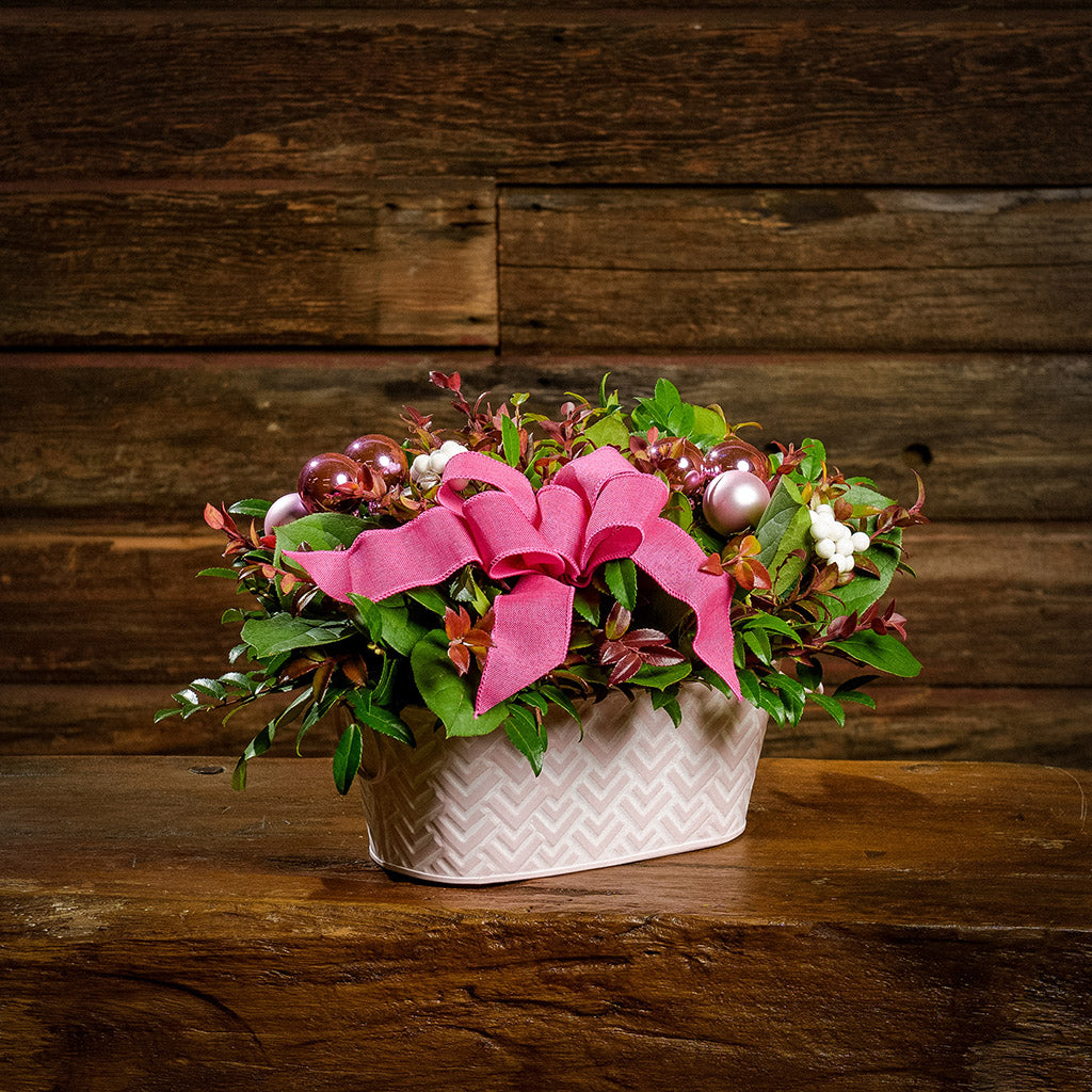 A centerpiece made of fresh salal, green huckleberry, red huckleberry, sweet huckleberry with pink ball clusters, white berries, and a magenta pink bow in a pink and white metal container sitting on a dark wooden bench.