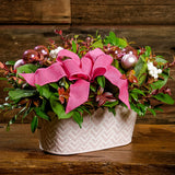 A close-up of a centerpiece made of fresh salal, green huckleberry, red huckleberry, sweet huckleberry with pink ball clusters, white berries, and a magenta pink bow in a pink and white metal container sitting on a dark wooden bench.