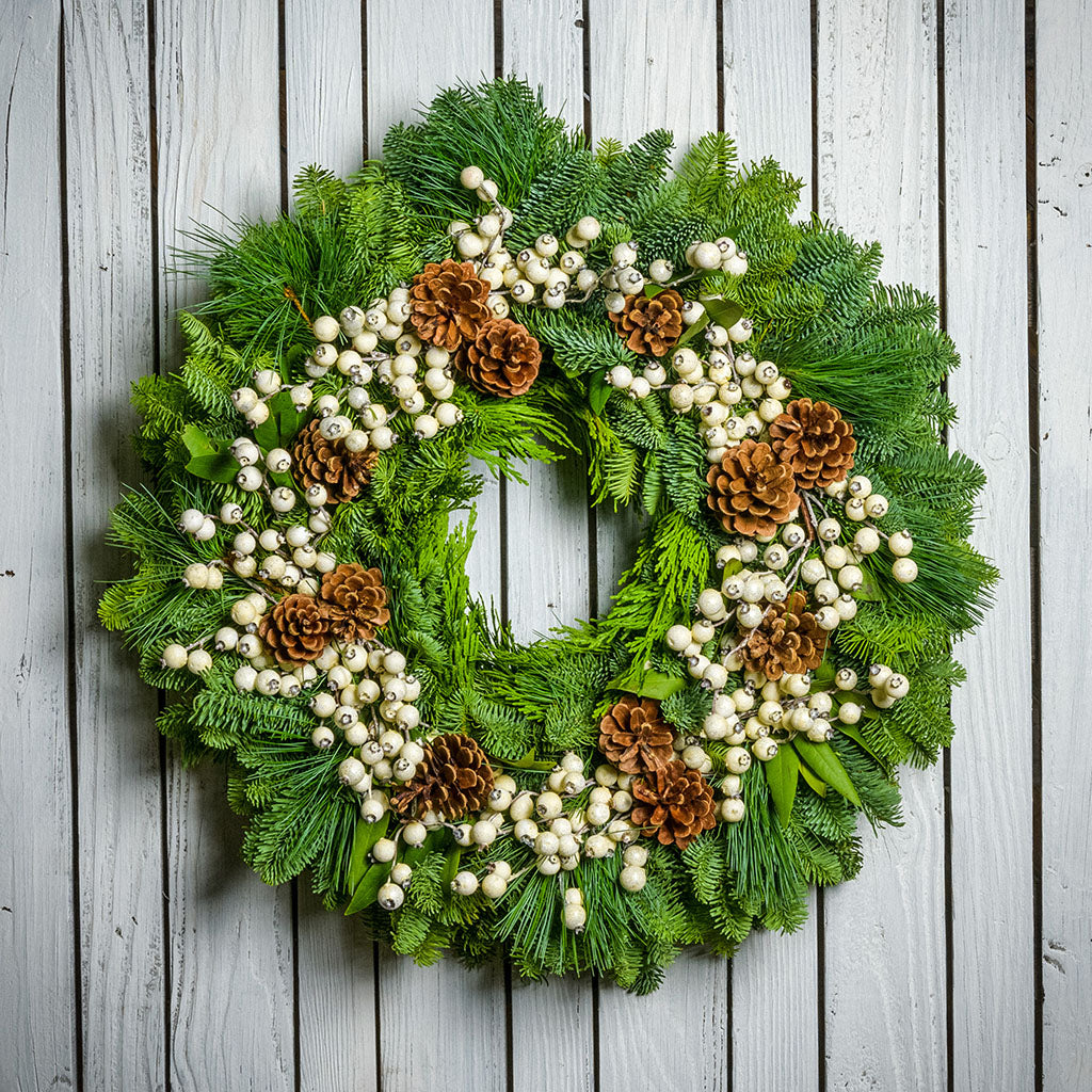 Holiday wreath made of noble fir, incense cedar, white pine, and bay leaf with faux white berries and leaves, and Australian pine cones hanging on a white fence