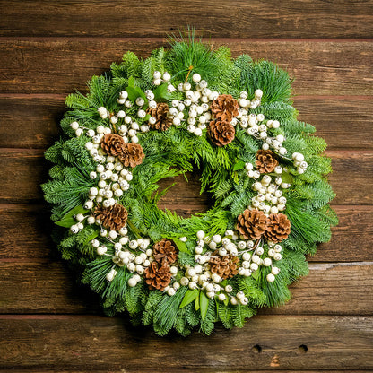 Holiday wreath made of noble fir, incense cedar, white pine, and bay leaf with faux white berries and leaves, and Australian pine cones hanging on a wooden wall