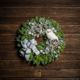 Holiday wreath of noble fir and western red cedar with a snowy white owl, faux flocked branch, small glitter fans, frosted branches, red berry clusters, shiny silver ball clusters, pinecones, and a silver glitter bow on a wood background.