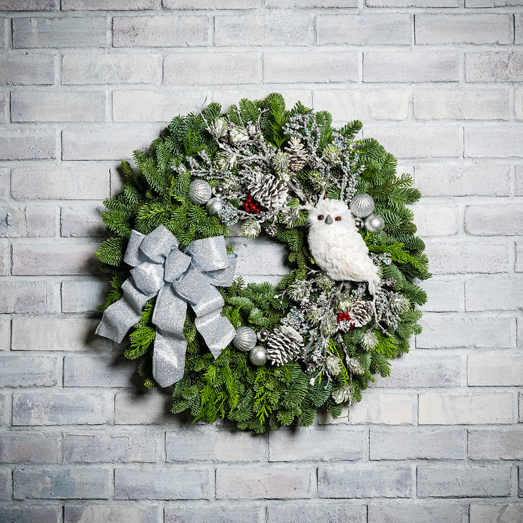 Holiday wreath of noble fir and western red cedar with a snowy white owl, faux flocked branch, small glitter fans, frosted branches, red berry clusters, shiny silver ball clusters, pinecones, and a silver glitter bow on a white brick background.