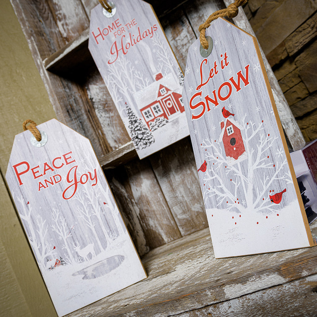 Set of 3 farmhouse grey-washed finish signs made of sturdy wood and hangs from a thick rustic jute rope. One sign says "Home for the Holidays" with red barn in a wintery holiday snow scene accented with trees. A second sign says "Peace and Joy" with snowy trees, birds, and a peaceful deer. The third sign says "Let it Snow" with cardinals birds visiting a sweet birdhouse all sitting on a shelf.