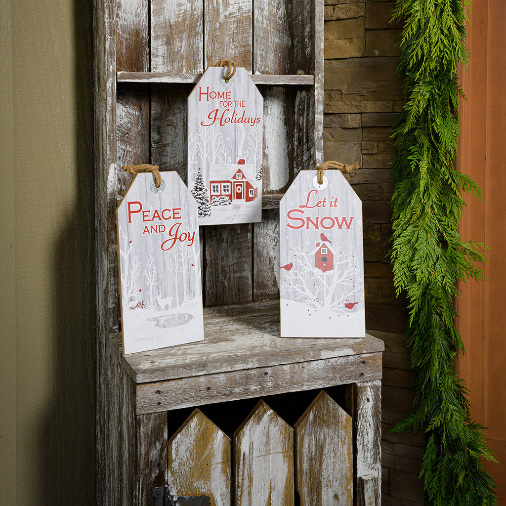 Set of 3 farmhouse grey-washed finish signs made of sturdy wood and hangs from a thick rustic jute rope. One sign says "Home for the Holidays" with red barn in a wintery holiday snow scene accented with trees. A second sign says "Peace and Joy" with snowy trees, birds, and a peaceful deer. The third sign says "Let it Snow" with cardinals birds visiting a sweet birdhouse all sitting on a shelf on a front porch, paired with garland.