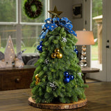 A table top tree made of noble fir with 4 gold ball clusters, 4 blue ball clusters, 8 silver berry clusters, a gold star tree-topper, and a blue starry bow sitting on a wood round on a table. 