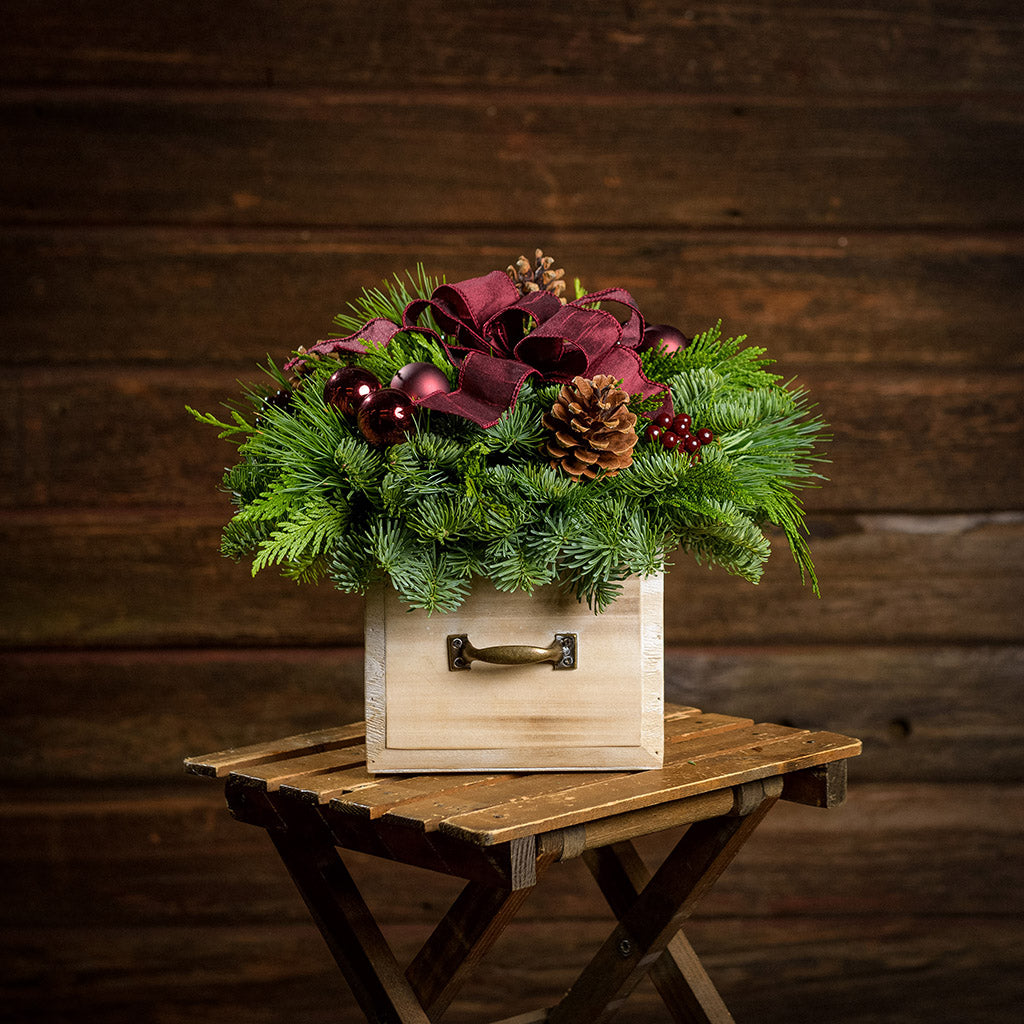 An arrangement made of noble fir, incense cedar, and white pine with Austrian pinecones, burgundy berry clusters, burgundy ball clusters, and a burgundy bow in a wood box with a metal handle with a dark wood background.