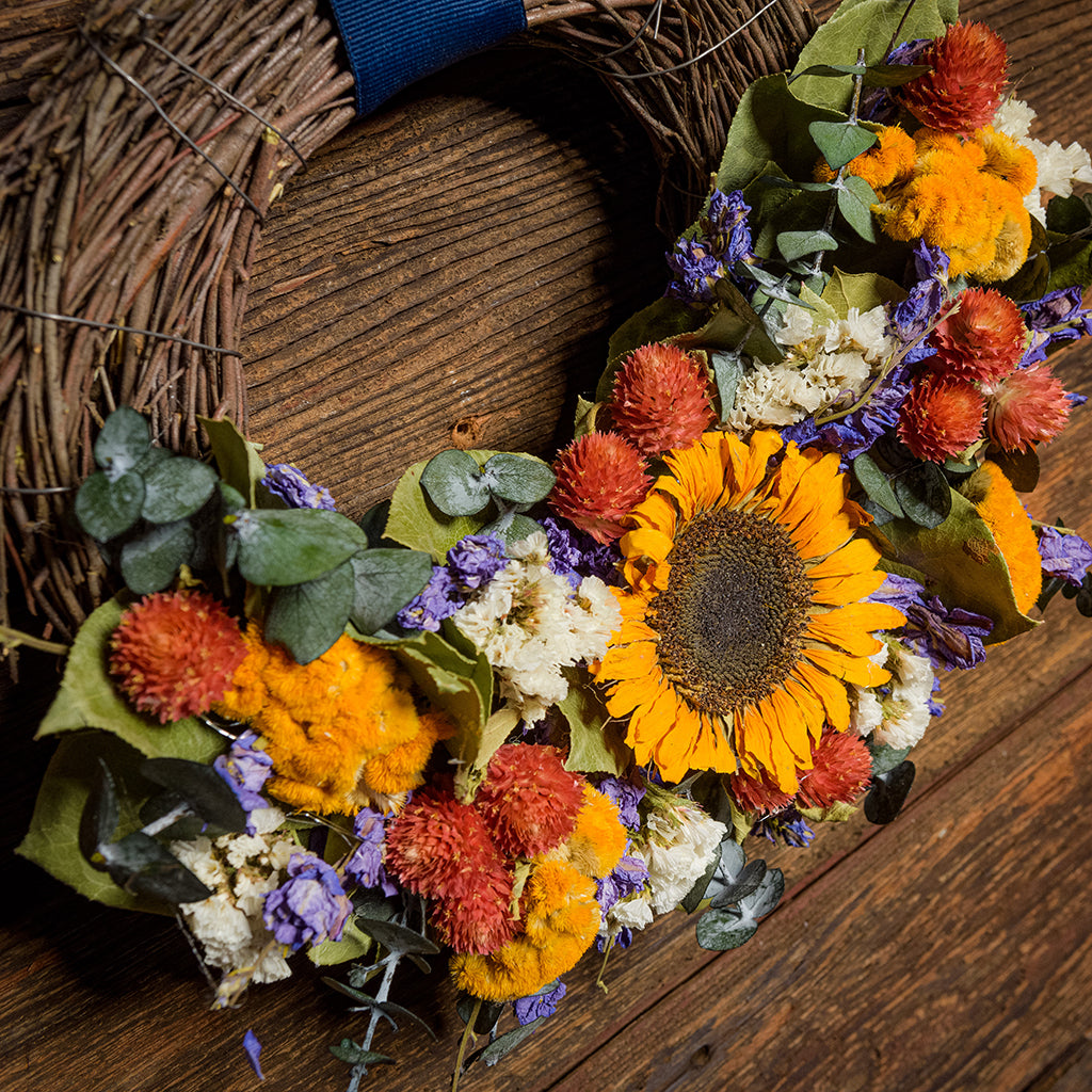 10" wreath made of eucalyptus, yellow coxcomb, white statice, red globe amaranthus, natural salal leaves, and real sunflowers hung on a dark wood background with grosgrain navy-blue bows.
