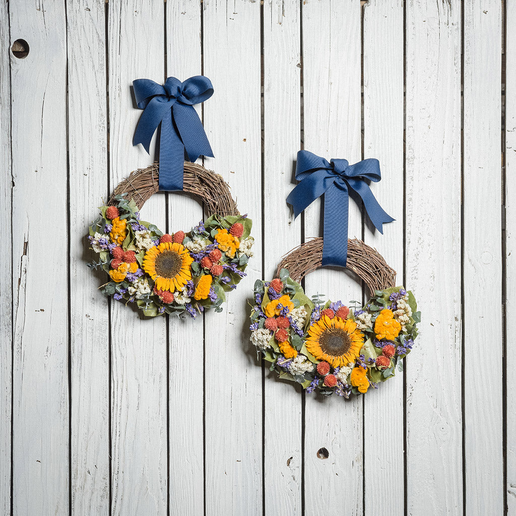 Two 10" wreaths made of eucalyptus, yellow coxcomb, white statice, red globe amaranthus, natural salal leaves, and real sunflowers hung on a white wood fence background with grosgrain navy-blue bows.