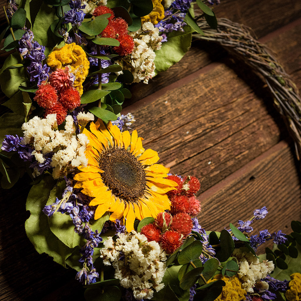 A wreath made of Green preserved eucalyptus, yellow coxcomb, white statice, larkspur, red globe amaranthus, natural salal leaves, and a sunflower on a dark wood background.