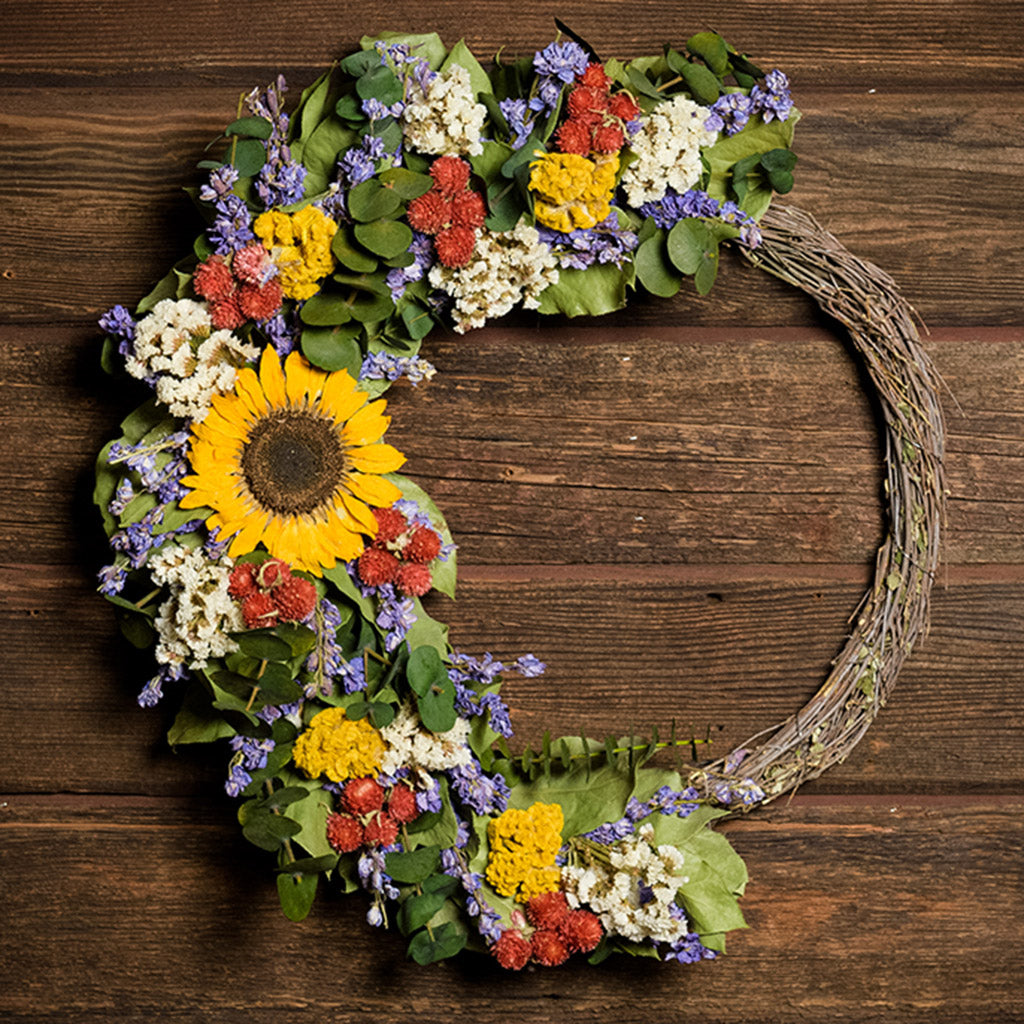 A wreath made of Green preserved eucalyptus, yellow coxcomb, white statice, larkspur, red globe amaranthus, natural salal leaves, and a sunflower on a dark wood background.