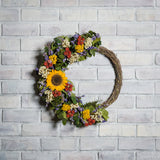 A wreath made of Green preserved eucalyptus, yellow coxcomb, white statice, larkspur, red globe amaranthus, natural salal leaves, and a sunflower on a with brick background.