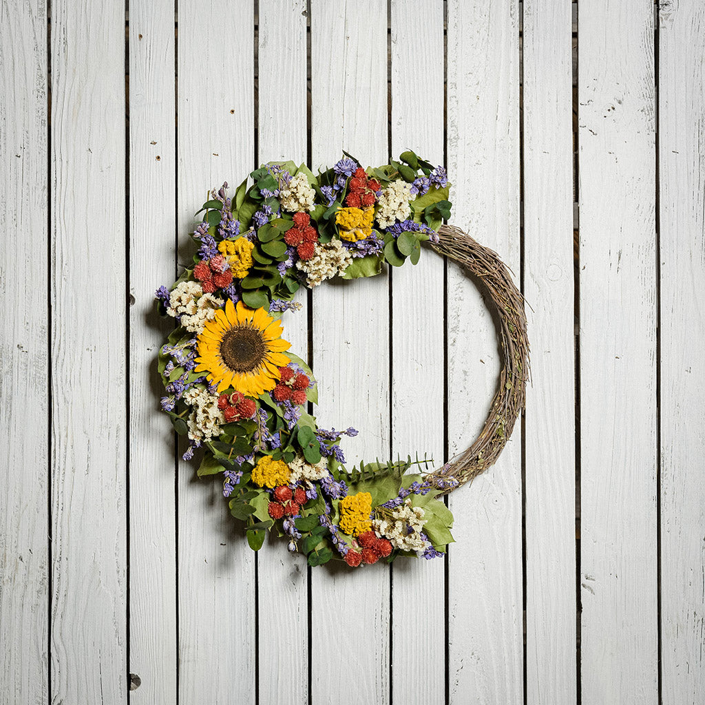 A wreath made of Green preserved eucalyptus, yellow coxcomb, white statice, larkspur, red globe amaranthus, natural salal leaves, and a sunflower on a white wood background.