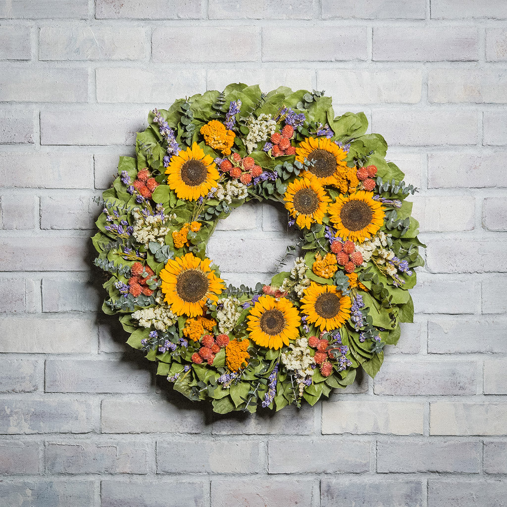 22" floral wreath made of  eucalyptus, yellow coxcomb, white statice, red globe amaranthus, salal leaves, and real sunflowers on a white brick background.