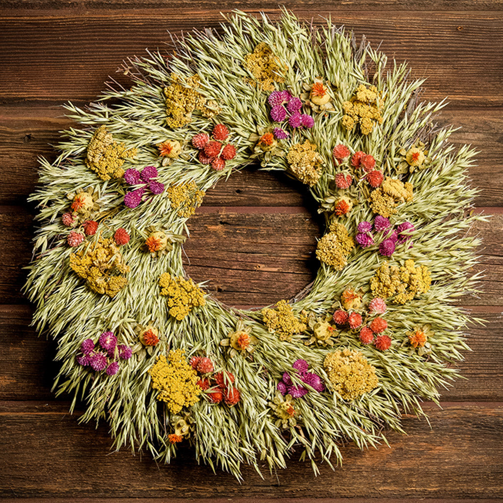 A wreath made of All-natural dried yarrow, natural oats, red and purple globe amaranthus, and safflowers on a dark wood background.