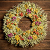 A wreath made of All-natural dried yarrow, natural oats, red and purple globe amaranthus, and safflowers on a dark wood background.