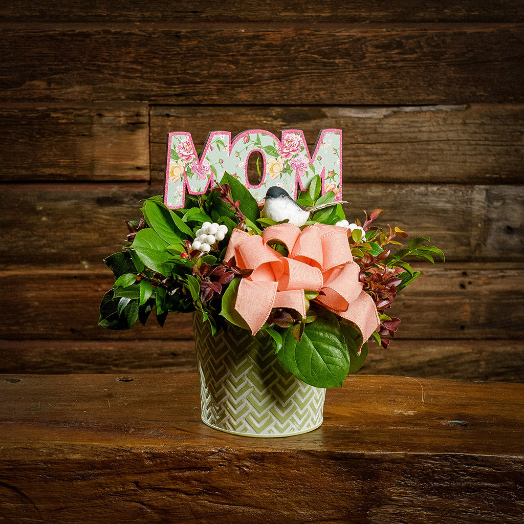 A centerpiece made of fresh salal, green huckleberry, and red huckleberry with white berries, reindeer moss, a peach bow, and a pink and green wood "Mom" sign in a green and white metal container sitting on a dark wooden bench.