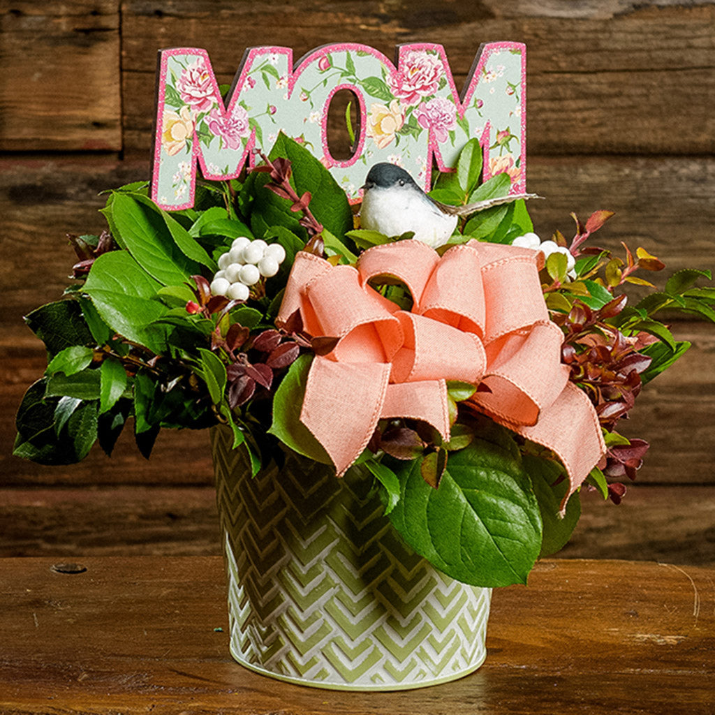 A close-up of a centerpiece made of fresh salal, green huckleberry, and red huckleberry with white berries, reindeer moss, a peach bow, and a pink and green wood "Mom" sign in a green and white metal container sitting on a dark wooden bench.