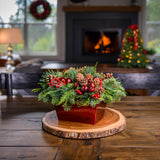 Centerpiece made of noble fir, cedar, and pine with Australian pine cones, red berry clusters, apple/pine cone picks, and a red metallic container on a wooden table