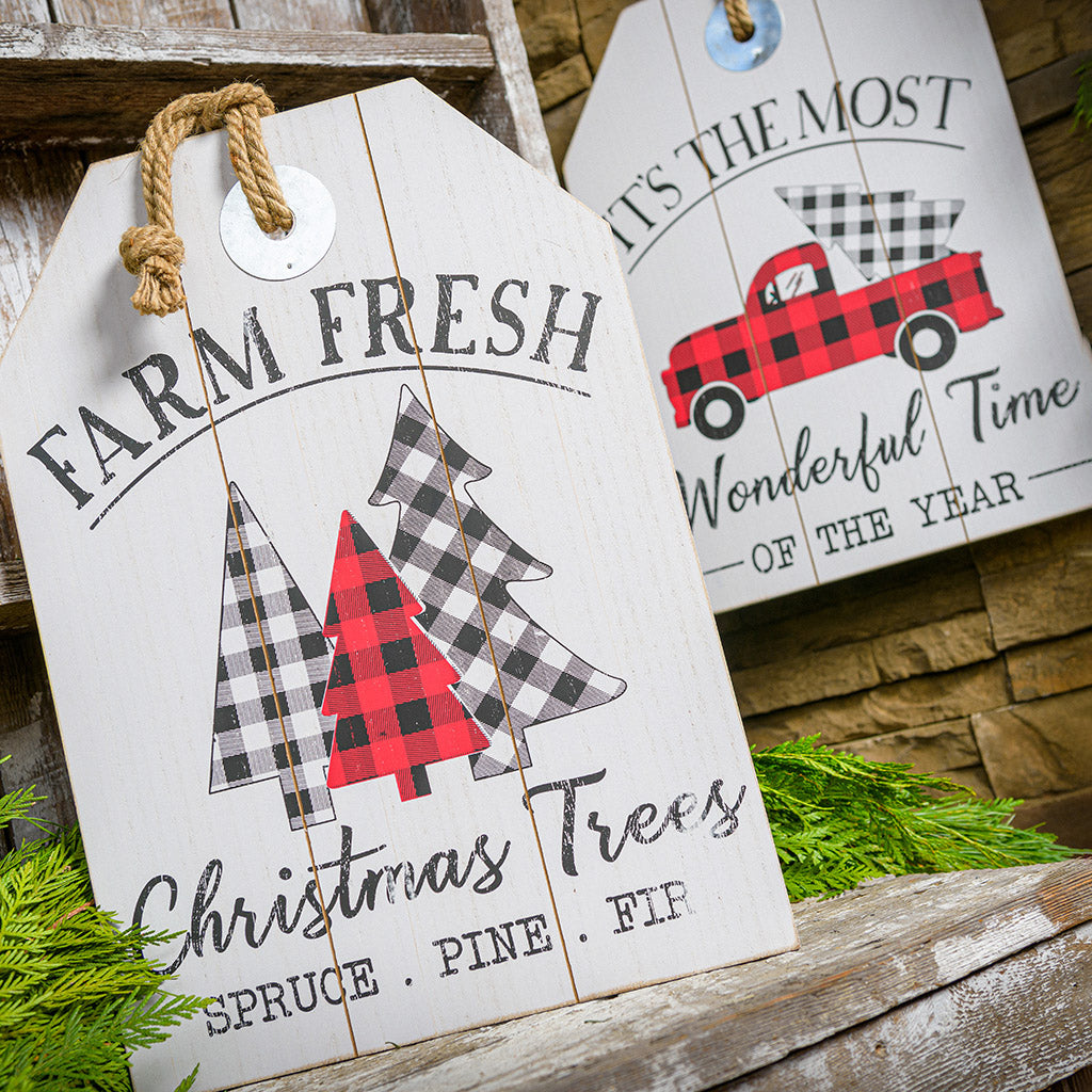 Sturdy wood signs hanging from a thick rustic jute rope. Both have beautiful rustic white-washed coloring. One sign says "Farm Fresh Christmas Trees - Spruce, Pine, Fir" with charming buffalo-plaid trees. The other sign says "It's the Most Wonderful Time of the Year" with a buffalo-plaid pickup truck bringing home a Christmas tree.