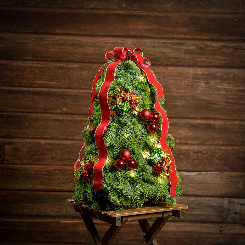 A tree made of noble fir, festive gift box decorations, red ball clusters, a red velvet bow with gold edging, and a strand of warm white lights with a dark wooden background.