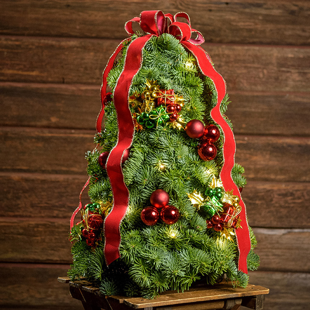 A tree made of noble fir, festive gift box decorations, red ball clusters, a red velvet bow with gold edging, and a strand of warm white lights with a dark wooden background.