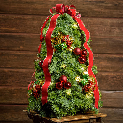 A tree made of noble fir, festive gift box decorations, red ball clusters, a red velvet bow with gold edging, and a strand of warm white lights with a dark wooden background.