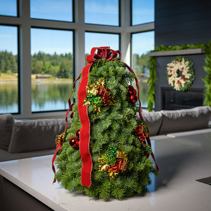 A tree made of noble fir, festive gift box decorations, red ball clusters, a red velvet bow with gold edging, and a strand of warm white lights sitting on a counter.