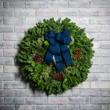 Christmas wreath made of fir, pine, cedar and juniper with pine cones and a blue brushed-linen bow on a light brick background.