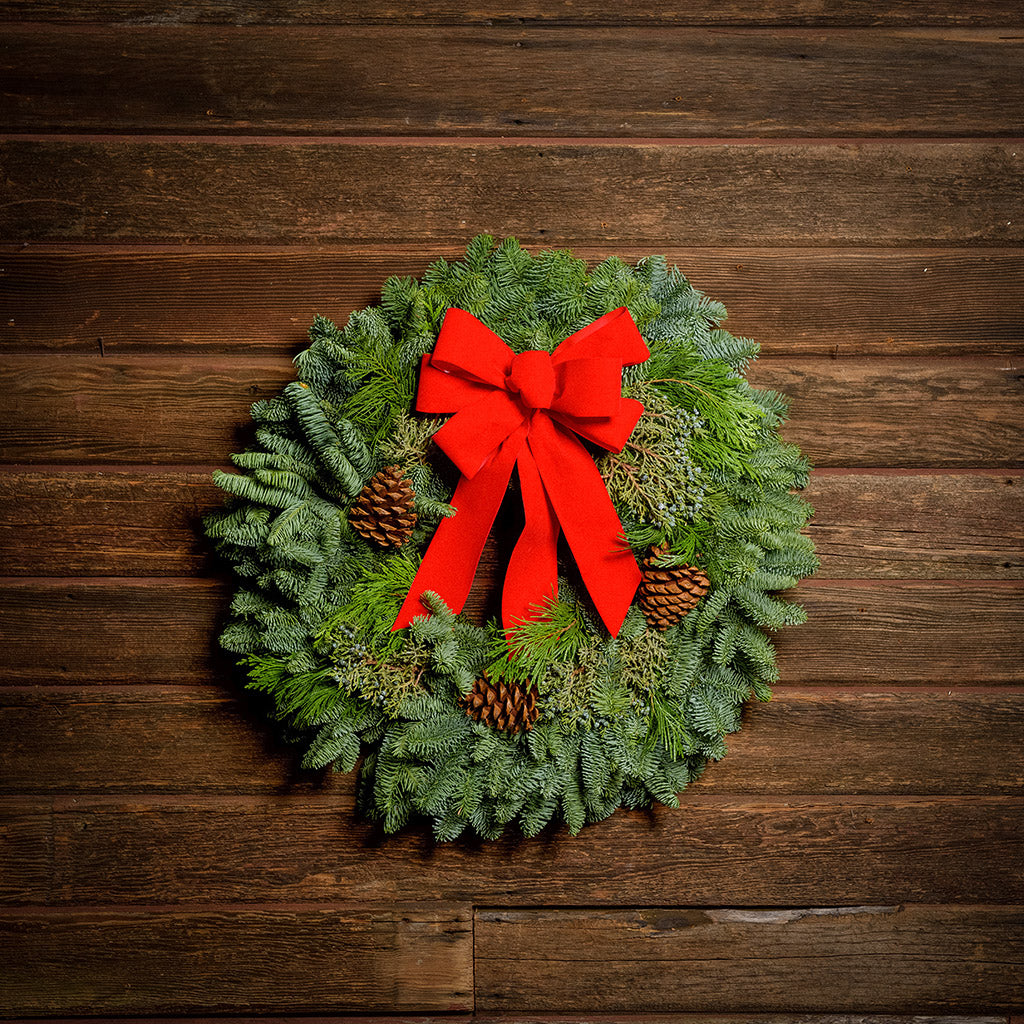 Christmas wreath made of fir, cedar, and juniper with pine cones and a bright holiday red bow on a wooden background.