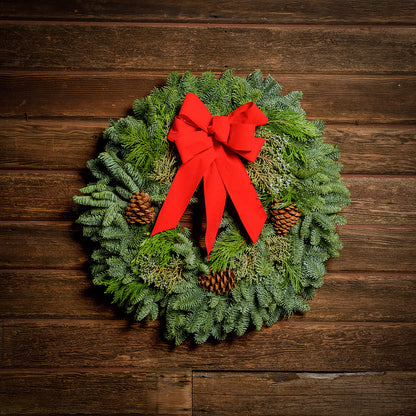 Christmas wreath made of fir, cedar, and juniper with pine cones and a bright holiday red bow on a wooden background.