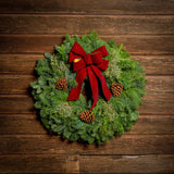 Christmas wreath made of fir, cedar, and juniper with pine cones and a burgundy with gold back bow on a light wooden background.