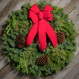 Christmas wreath made of fir, cedar, and juniper with pine cones and a bright holiday red bow on a light wooden background.