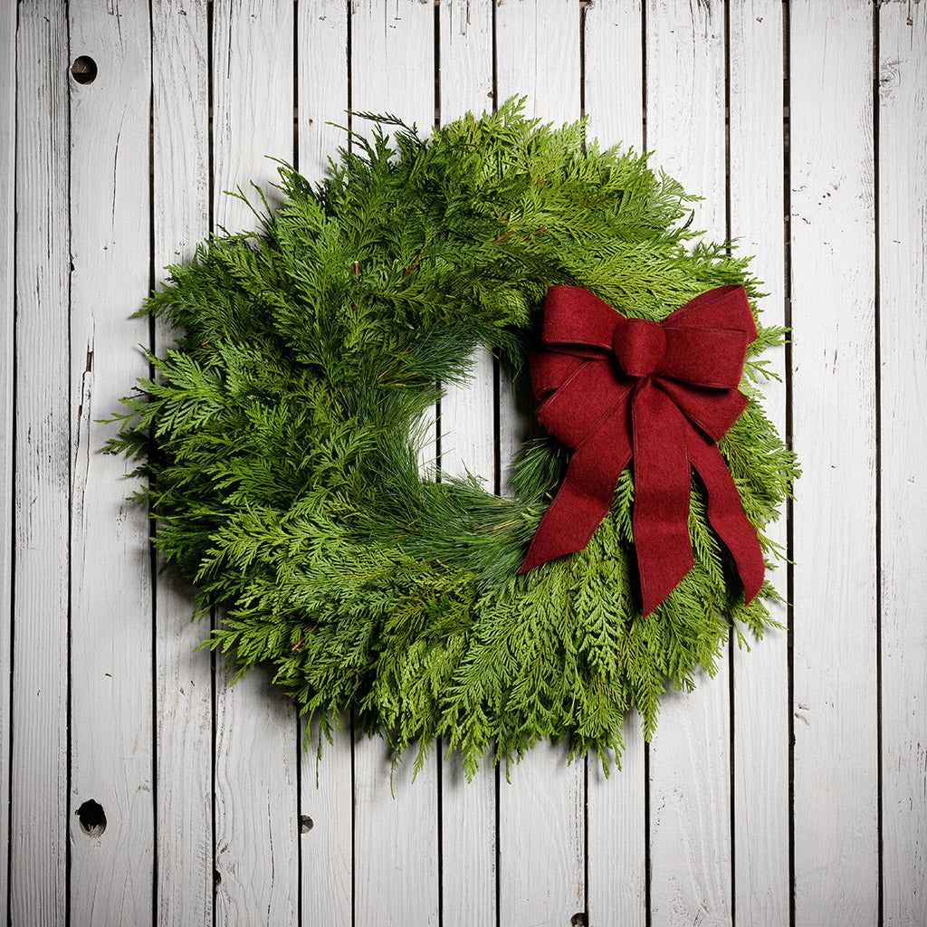 Fresh Evergreen Christmas wreath with cedar and pine and a brushed red bow on a white wood background.
