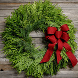 Fresh Evergreen Christmas wreath with cedar and pine and a brushed red bow on a light wood background.