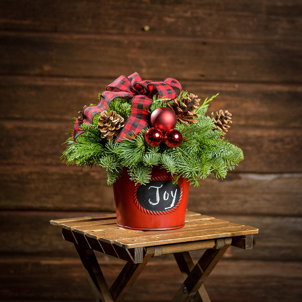 An arrangement made of noble fir, incense cedar, and white pine with Austrian pinecones, red balls, and a red and black plaid bow in a red metal container with a chalkboard (chalk not included) with a dark wooden background.