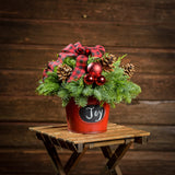 An arrangement made of noble fir, incense cedar, and white pine with Austrian pinecones, red balls, and a red and black plaid bow in a red metal container with a chalkboard (chalk not included) with a dark wooden background.