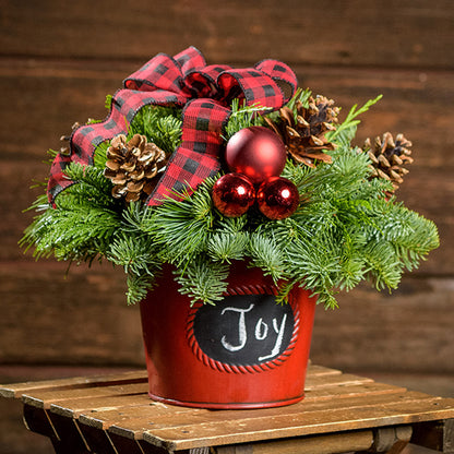 An arrangement made of noble fir, incense cedar, and white pine with Austrian pinecones, red balls, and a red and black plaid bow in a red metal container with a chalkboard (chalk not included) with a dark wooden background.