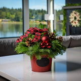 An arrangement made of noble fir, incense cedar, and white pine with Austrian pinecones, red balls, and a red and black plaid bow in a red metal container with a chalkboard (chalk not included) sitting on a counter.