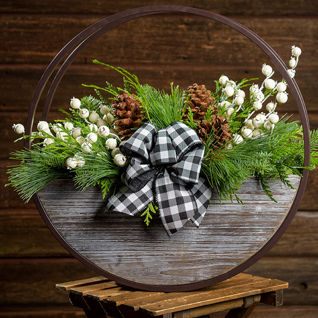 An arrangement made of noble fir, Western red cedar, and white pine with ponderosa pinecones, white mini pomegranate branches, and a black and white gingham linen bow in a wood and metal hanging container with a dark wood background.