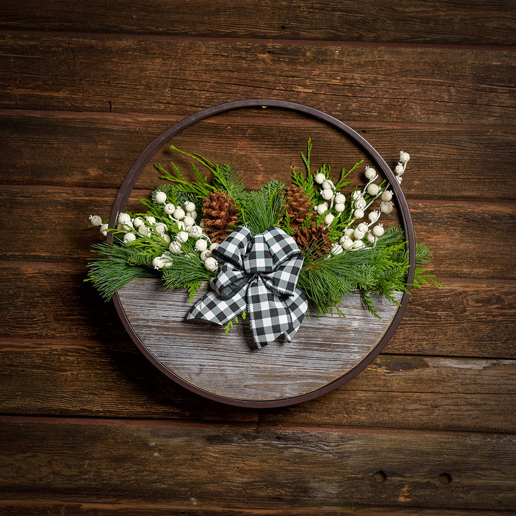 An arrangement made of noble fir, Western red cedar, and white pine with ponderosa pinecones, white mini pomegranate branches, and a black and white gingham linen bow in a wood and metal hanging container with a dark wood background.