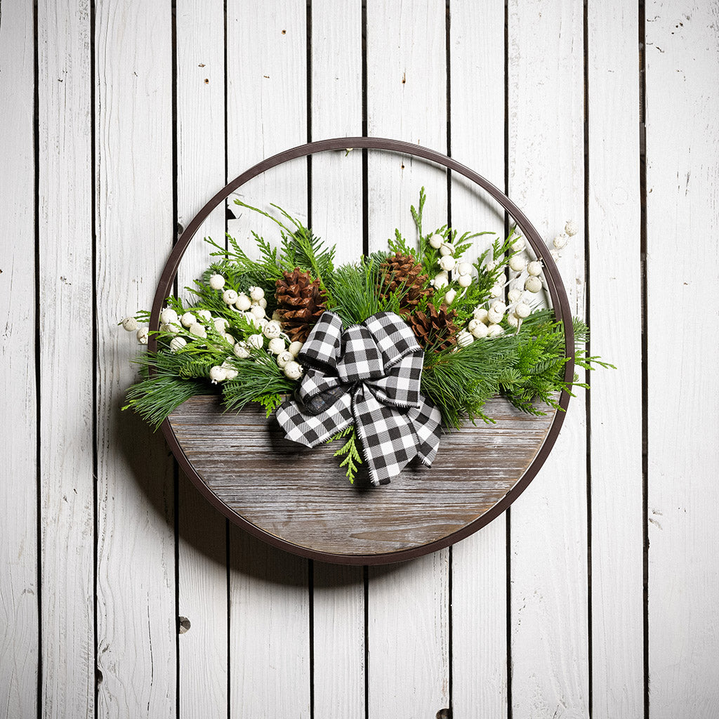 An arrangement made of noble fir, Western red cedar, and white pine with ponderosa pinecones, white mini pomegranate branches, and a black and white gingham linen bow in a wood and metal hanging container with a white wood background.