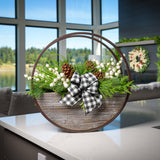 An arrangement made of noble fir, Western red cedar, and white pine with ponderosa pinecones, white mini pomegranate branches, and a black and white gingham linen bow in a wood and metal hanging container sitting on a counter. 