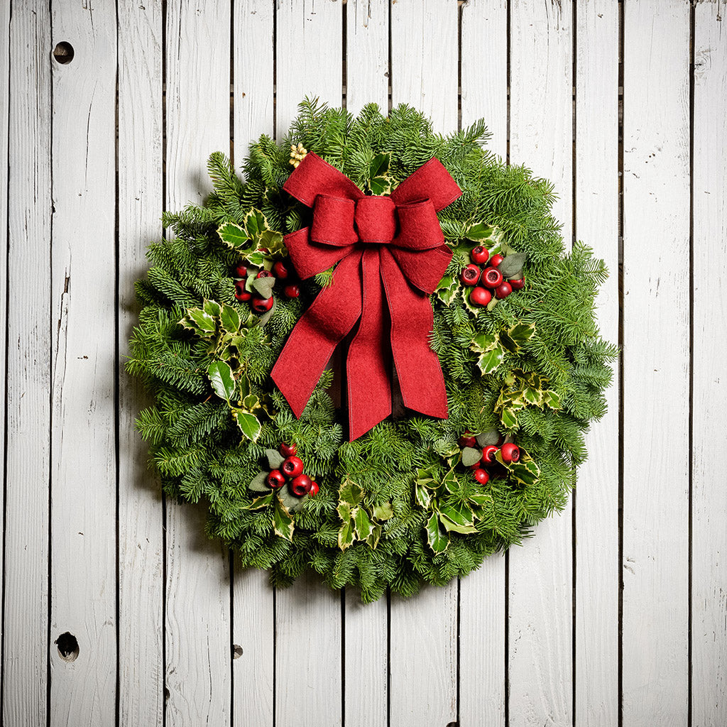 Christmas wreath made of noble fir and variegated holly with 4 country-berry clusters and a red brushed-linen bow on a white wood background.