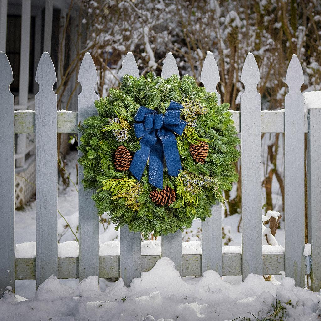 Christmas wreath made of fir, pine, cedar and juniper with pine cones and a blue brushed-linen bow on a white picket fence..