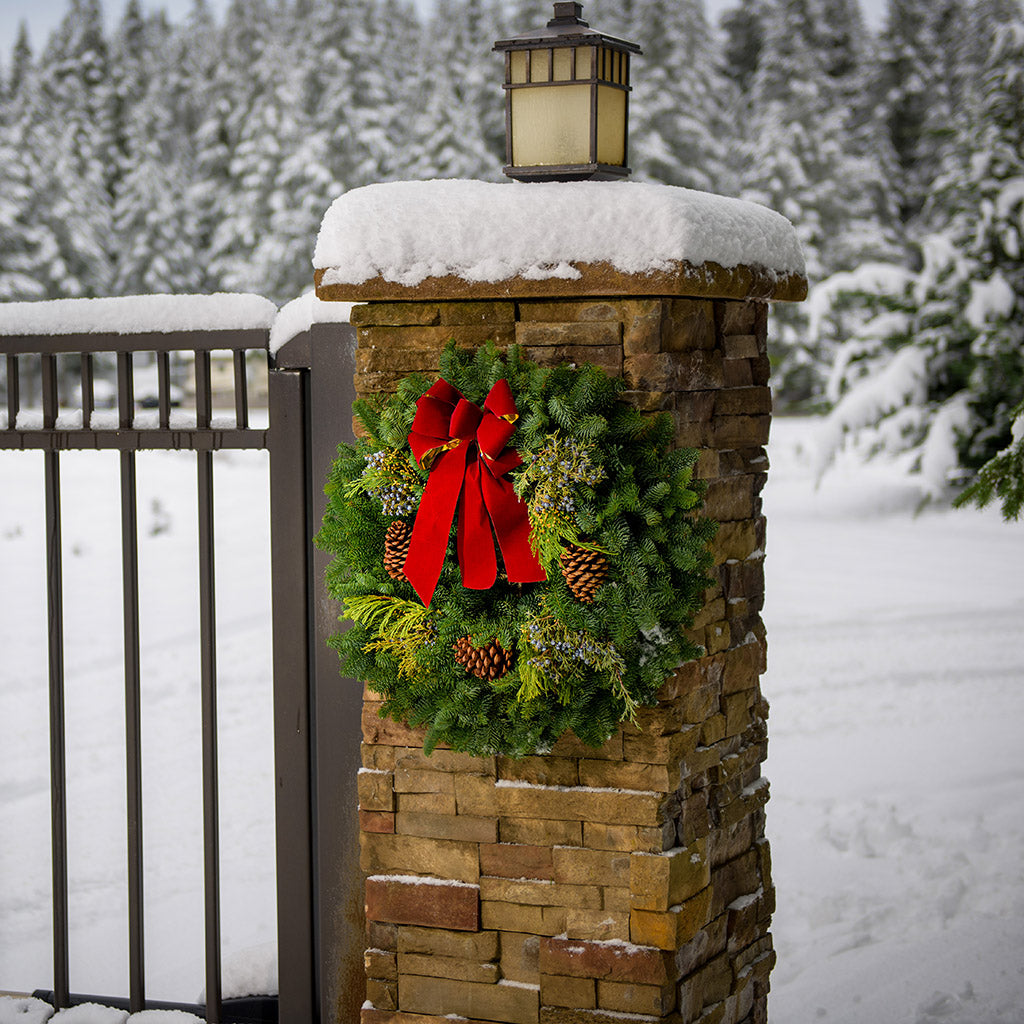 Christmas wreath made of fir, pine, cedar and juniper with pine cones and a gold-backed red velveteen bow hung on natural brick pillar.