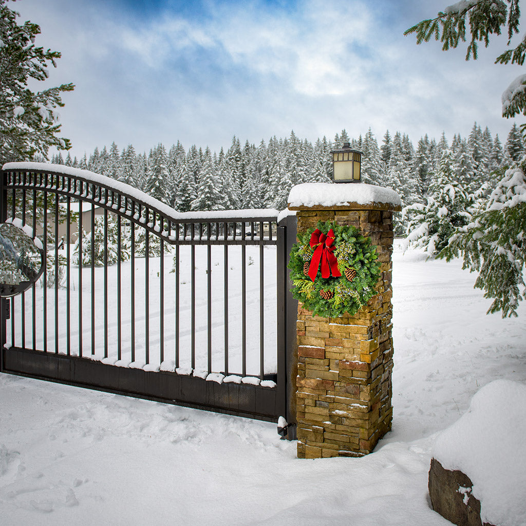 Christmas wreath made of fir, pine, cedar and juniper with pine cones and a gold-backed red velveteen bow hung on a natural brick pillar.