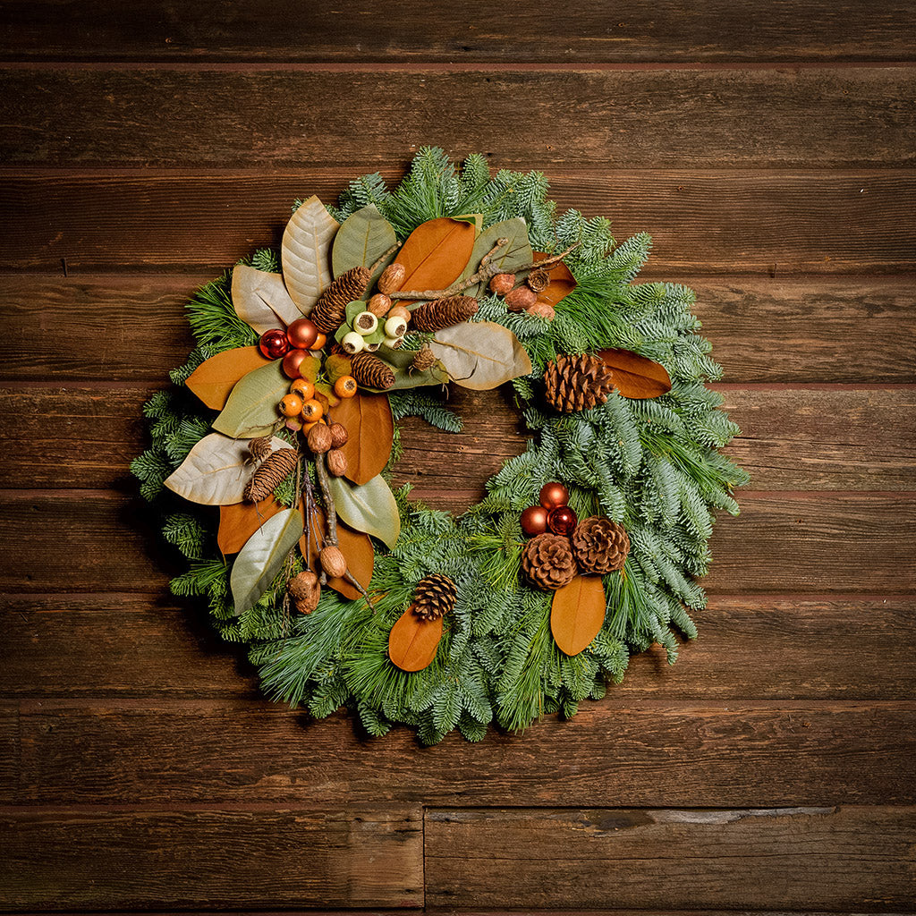 A wreath made of noble fir, white pine, and magnolia leaves with ponderosa and Austrian pinecones, copper balls, cream and orange seed pods, and branches of assorted nuts and cones on a dark wood background.