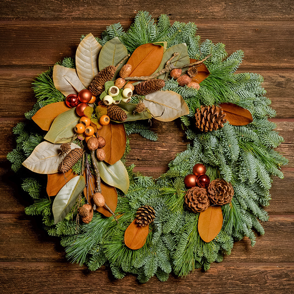A wreath made of noble fir, white pine, and magnolia leaves with ponderosa and Austrian pinecones, copper balls, cream and orange seed pods, and branches of assorted nuts and cones on a dark wood background.