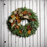A wreath made of noble fir, white pine, and magnolia leaves with ponderosa and Austrian pinecones, copper balls, cream and orange seed pods, and branches of assorted nuts and cones on a white wood background.