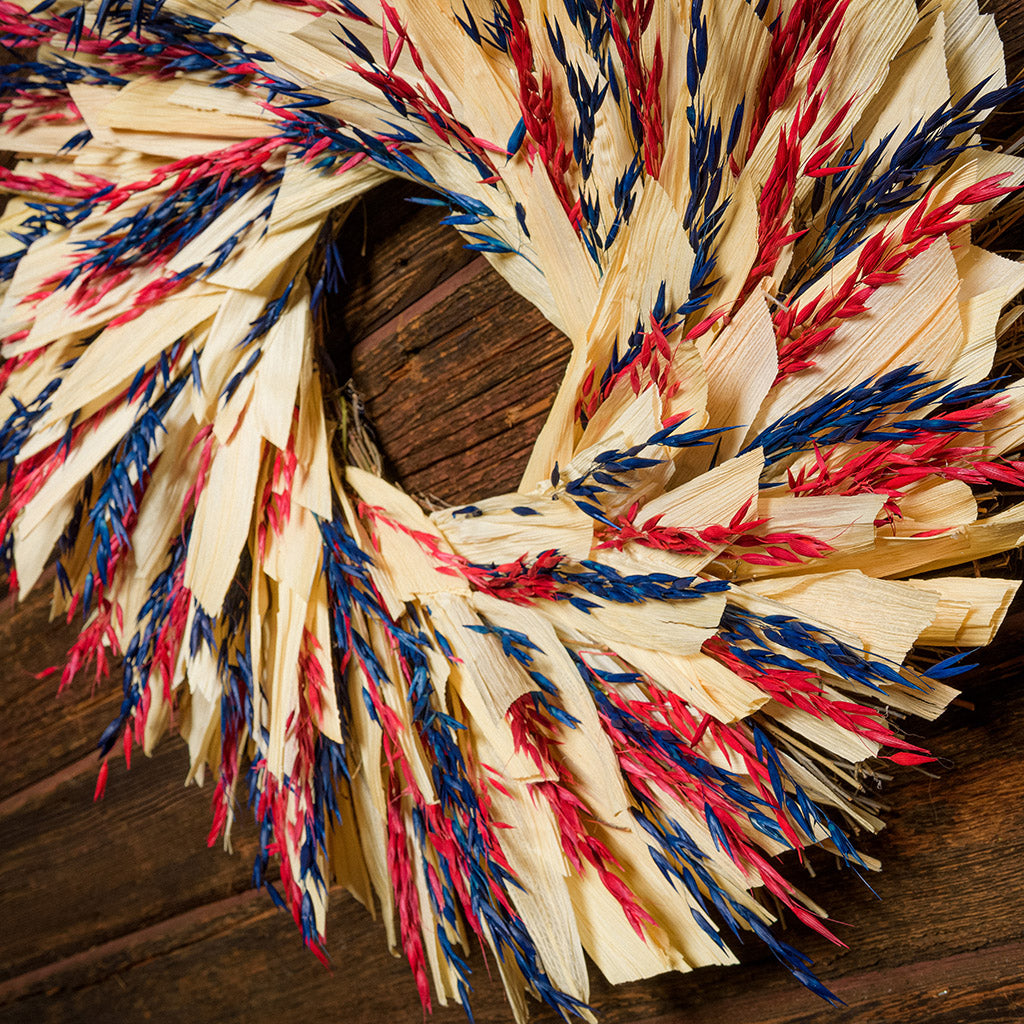 22" wreath made of red and blue oats and natural corn husk on a dark wood background.